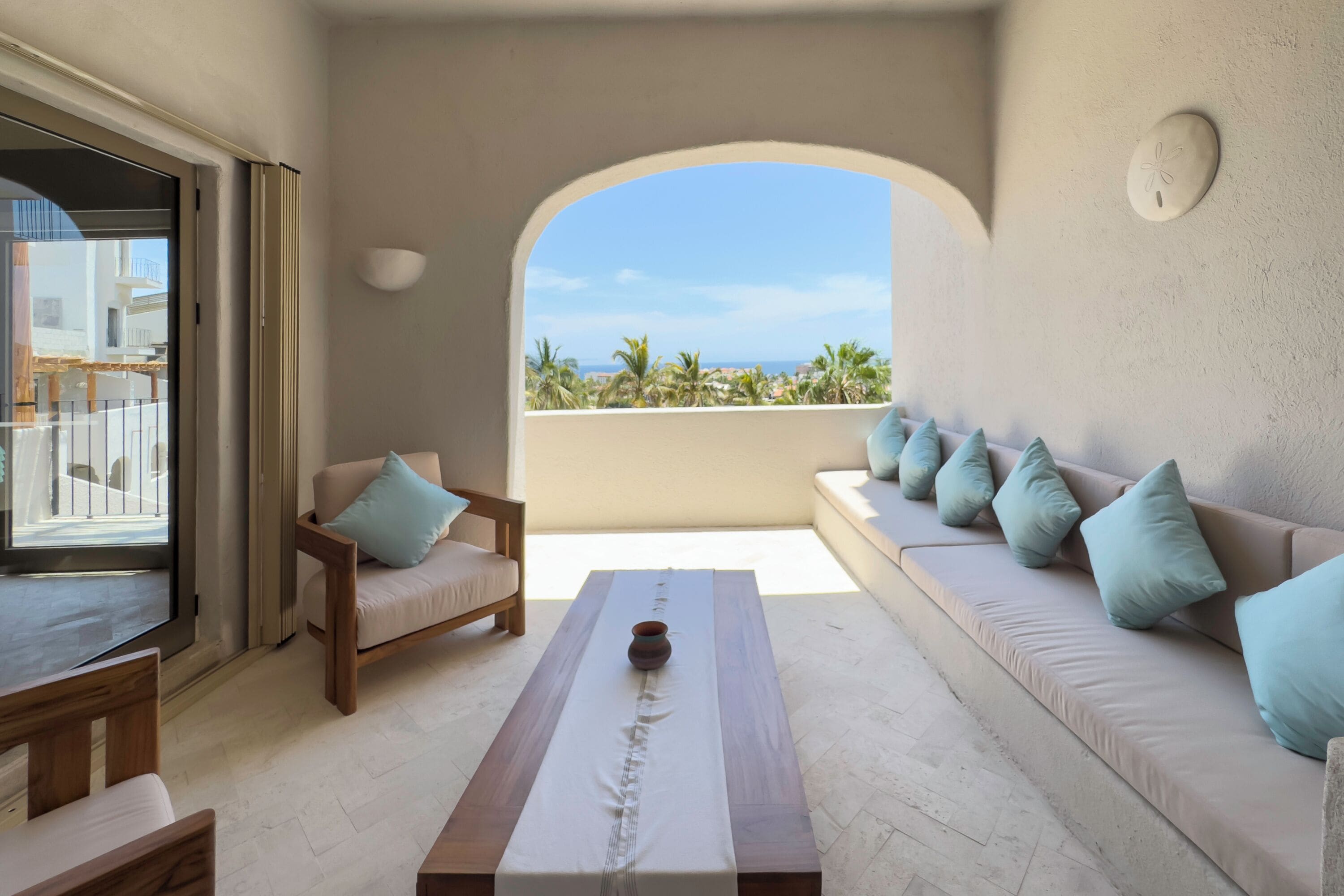 Un salon moderne avec un canapé, une table en bois et une vue sur des palmiers et une plage, visible à travers une arche.
