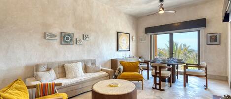 A spacious living area with a sofa, armchair, coffee table, dining table, and chairs. There are wall art pieces and a large window providing a view.