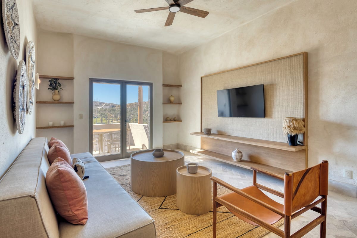 Una sala de estar moderna con un sofá, una silla de madera, un televisor montado en la pared y un ventanal con vistas al exterior.