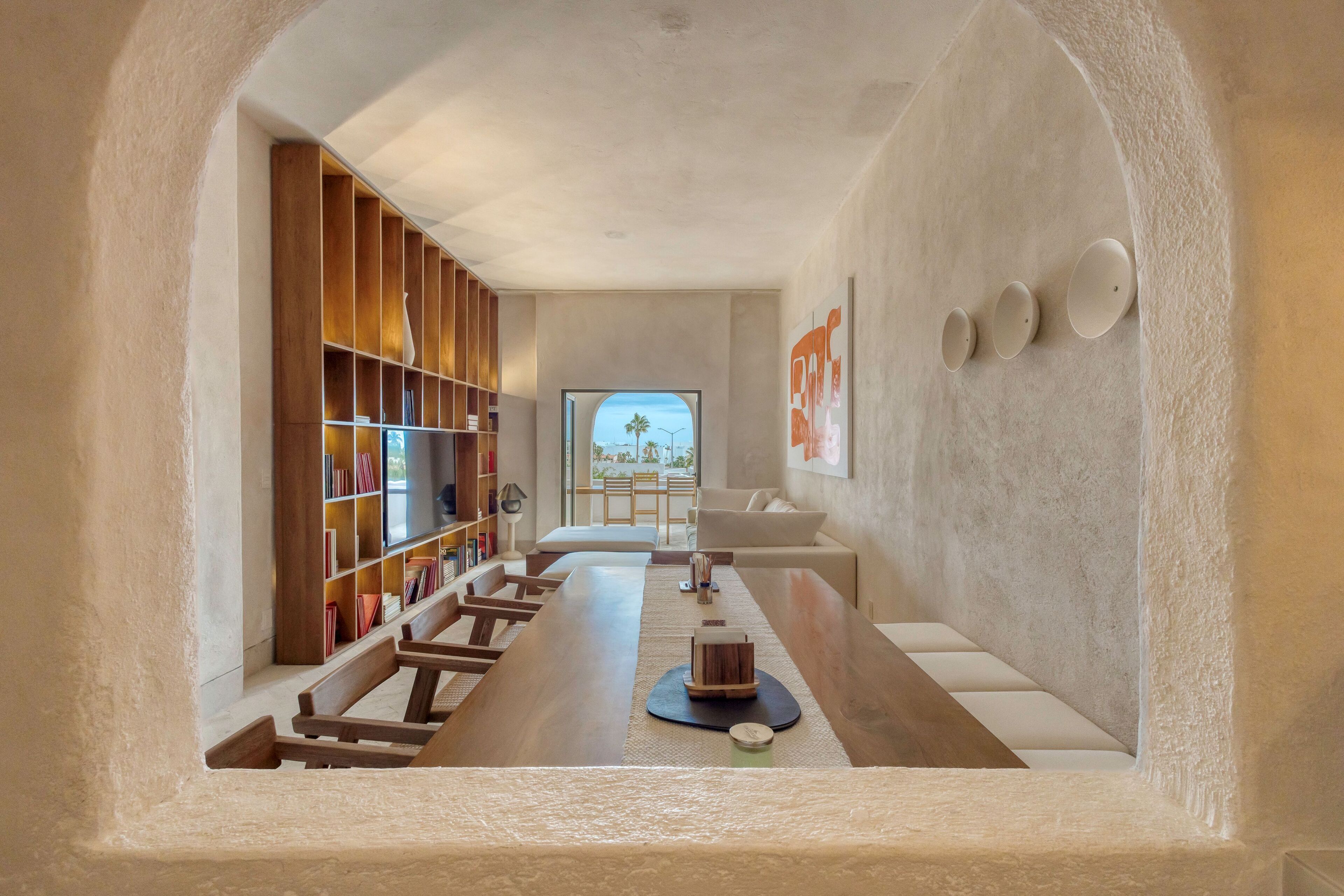 A modern interior with a long wooden table, bookshelves, and a view through a large window.