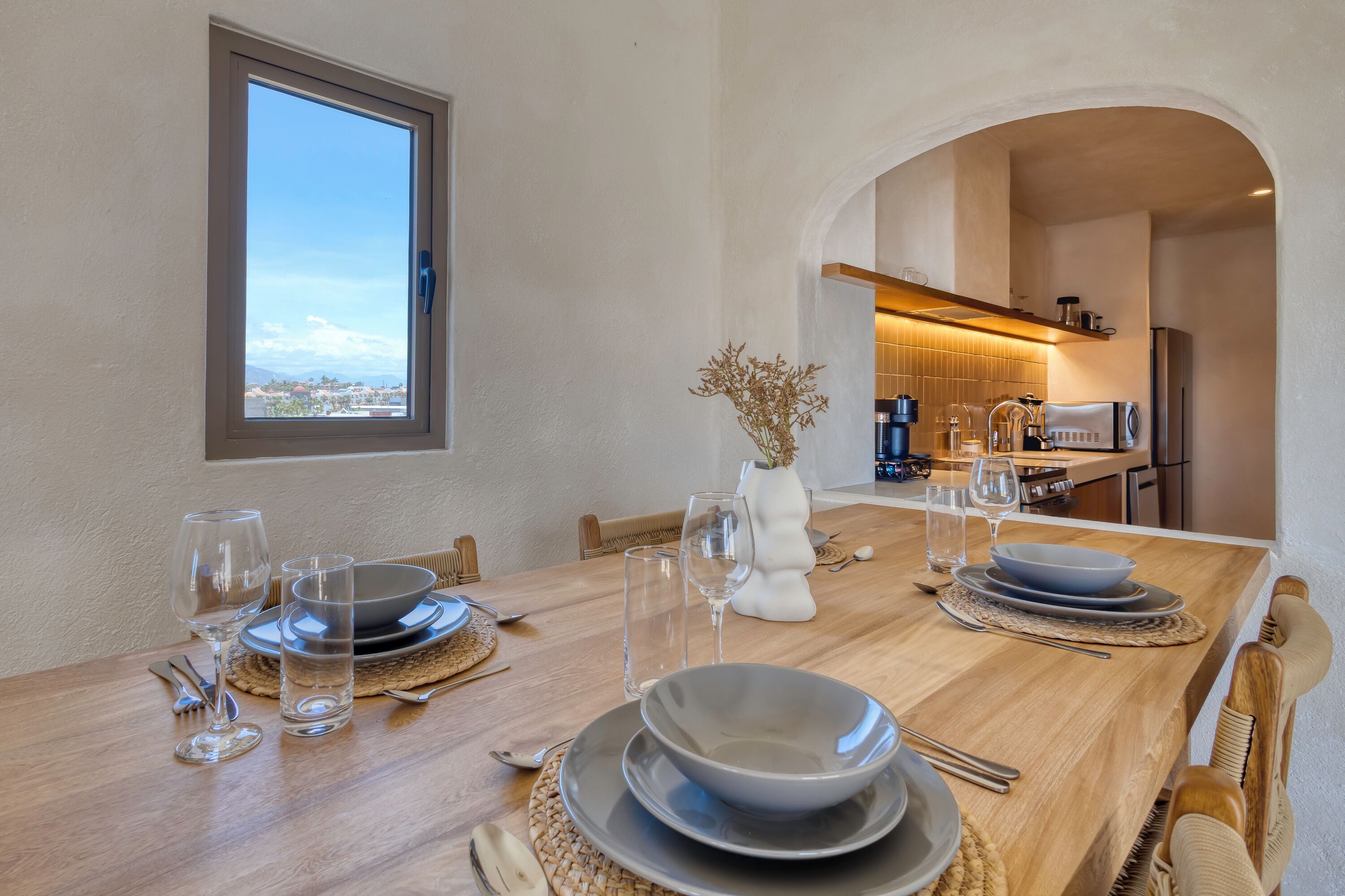 Una mesa de comedor dispuesta con platos, tazones, vasos y cubiertos, con vista a la ciudad a través de una ventana.