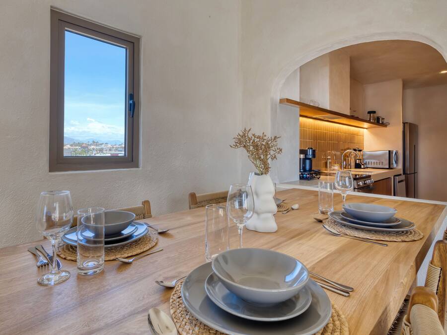 Una mesa de comedor dispuesta con platos, tazones, vasos y cubiertos, con vista a la ciudad a través de una ventana.