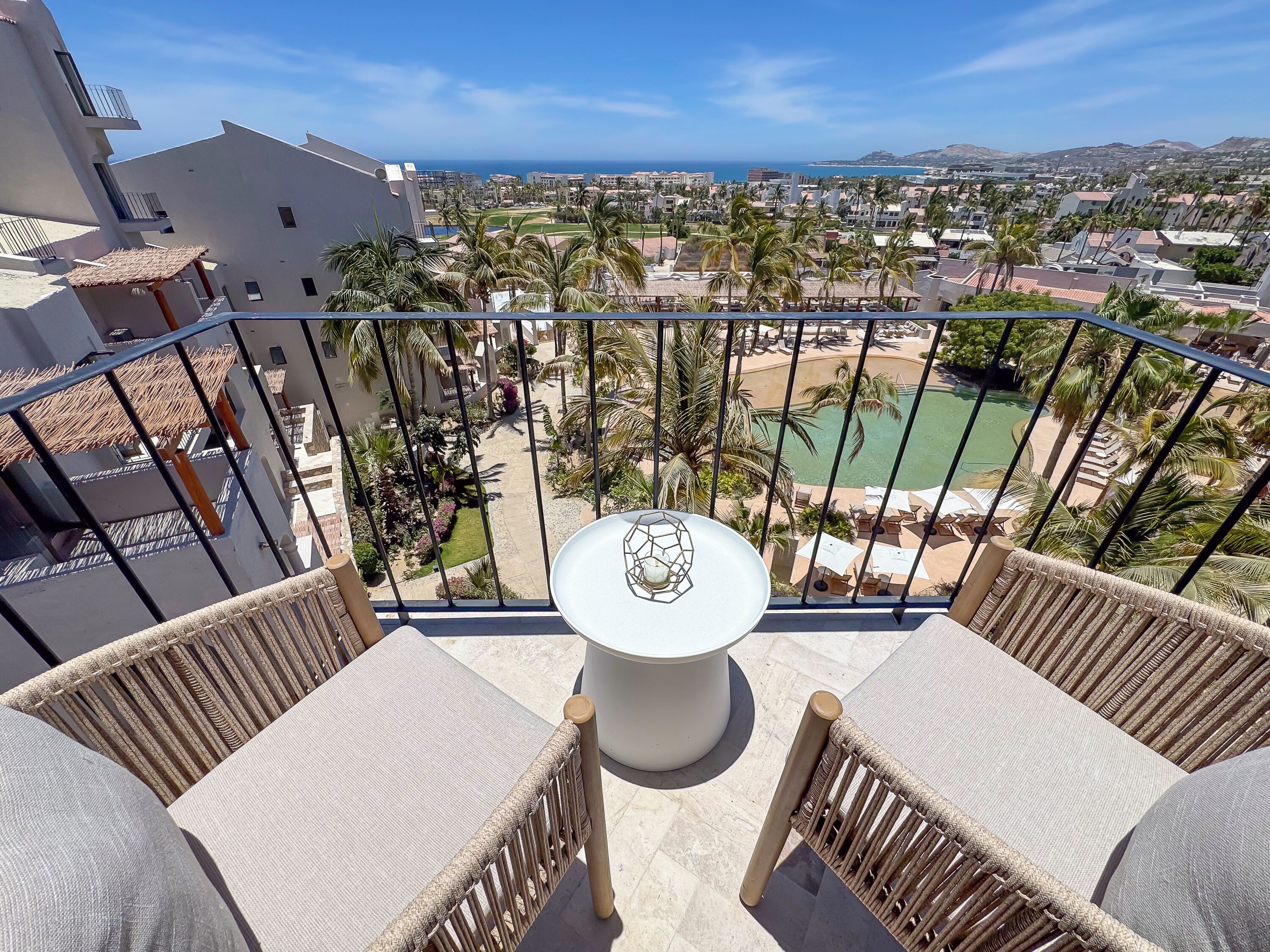 Ein Balkon mit Korbmöbeln, einem kleinen Tisch und Blick auf einen Ferienort mit Pool und Palmen.