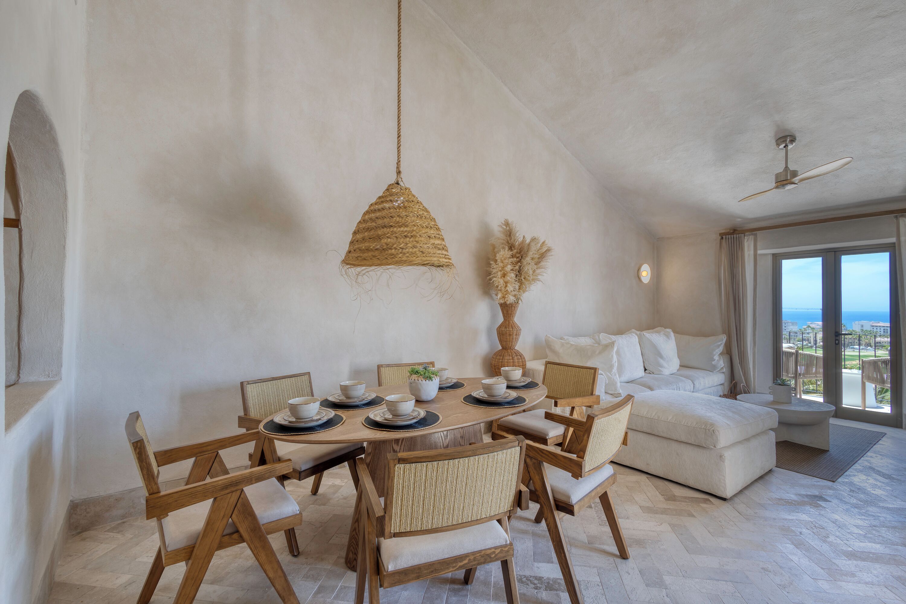 Un vaste espace de vie comprenant un coin repas avec une table pour quatre personnes, entourée de chaises en bois. Une grande fenêtre offre une vue sur l’océan.