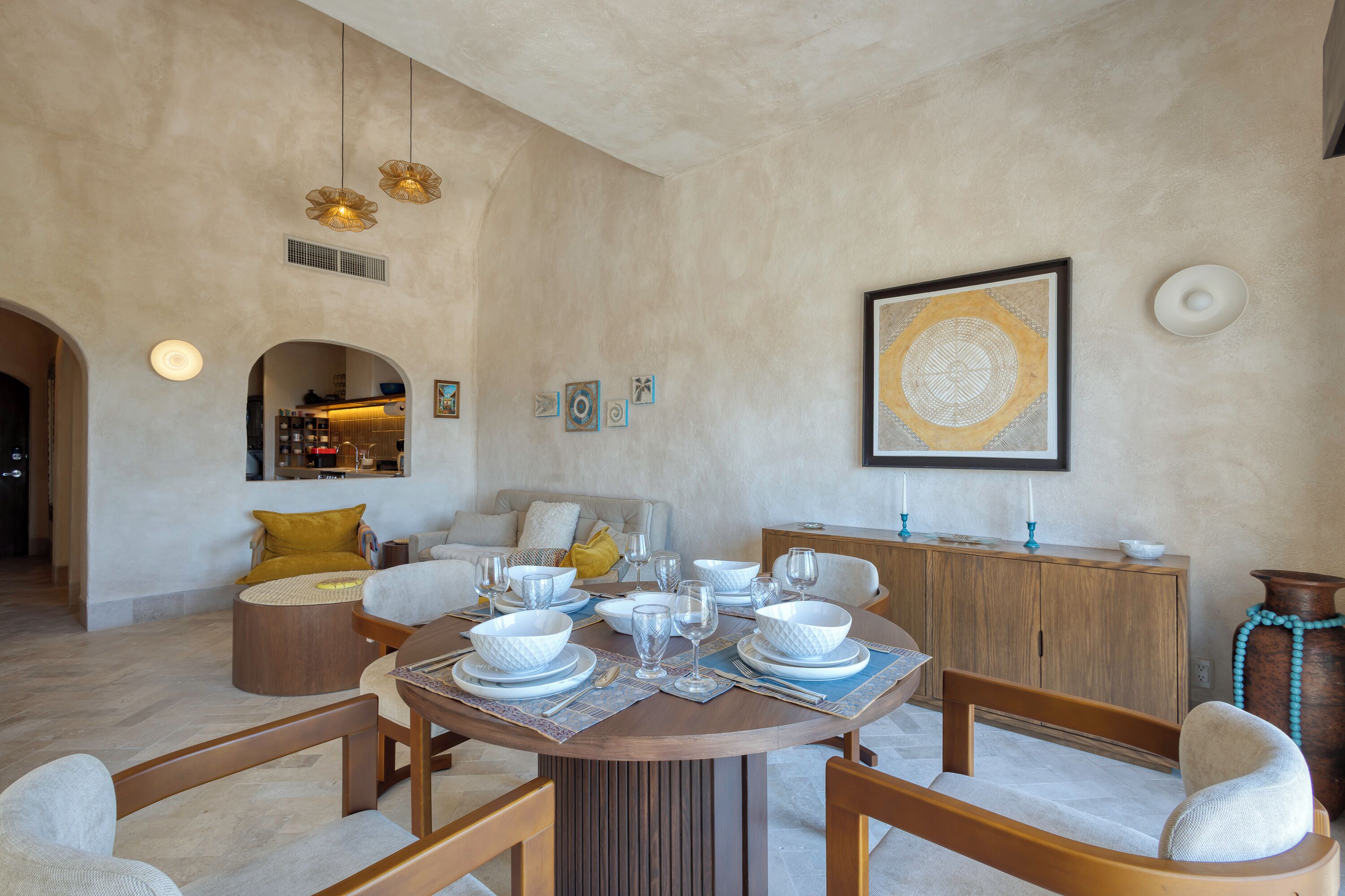 A dining area with a round table set for six, surrounded by wooden chairs. There is a wooden cabinet with decorative items on top, and a framed artwork on the wall.