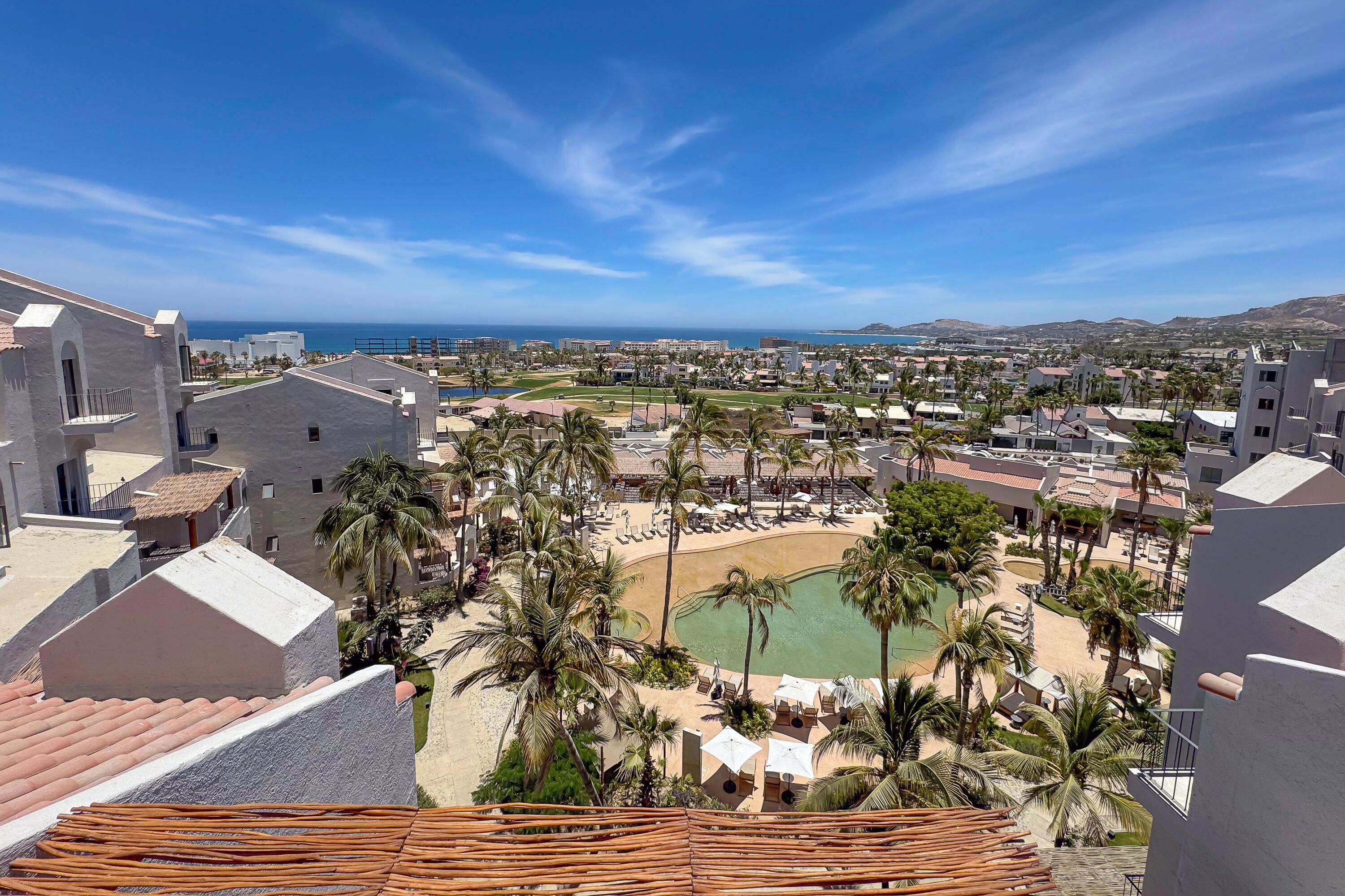 A resort with a pool surrounded by palm trees and lounge chairs, overlooking a beach and ocean.