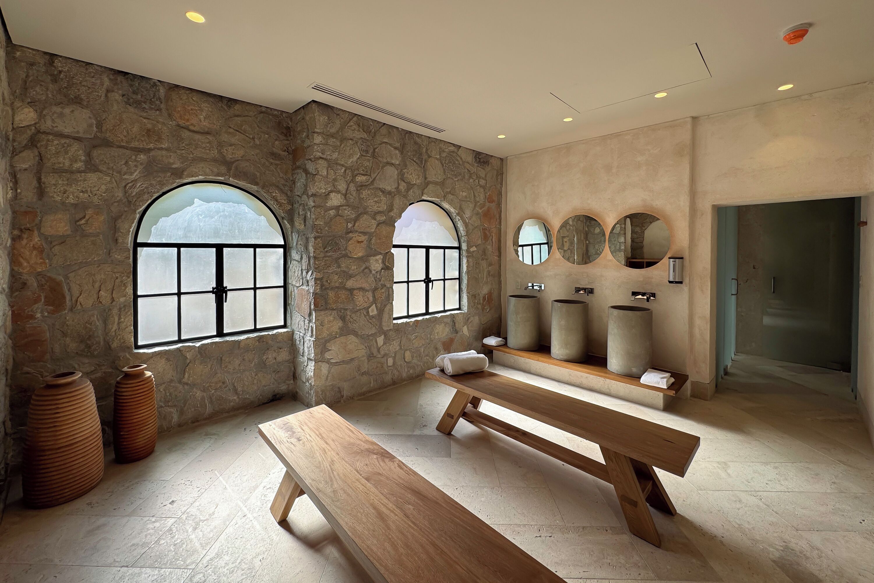 Una habitación con paredes de piedra, amplias ventanas de arco, bancos de madera y una chimenea de piedra.