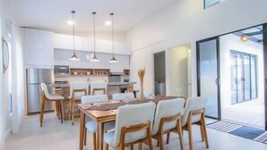 A modern kitchen with white cabinets, a dining table with chairs, pendant lights, and a view of the outdoors through a glass door.