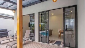 A modern outdoor patio with a wooden deck, a glass door leading to a pool area, and a dining table with chairs.