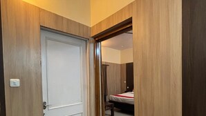 A hotel room with a wooden door, a bed with a red blanket, and a small trash bin. - Bagga Vilas (Jaipur)