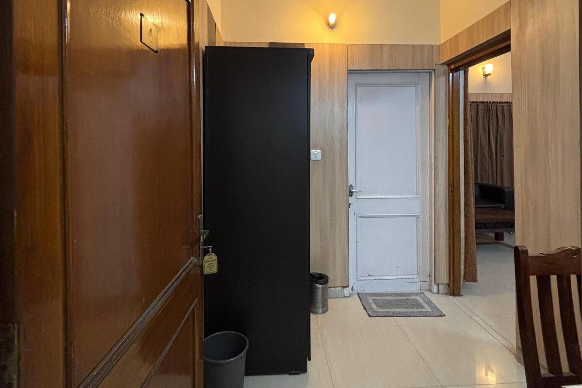 Un couloir avec une porte en bois, un réfrigérateur noir, une poubelle et une porte blanche au fond.