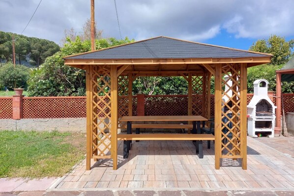 Un mirador de madera con un banco, rodeado por un patio de ladrillo y una cerca de enrejado rojo.
