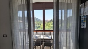 Un balcón con mesa y sillas, vistas a zonas verdes y edificios, y una ventana con cortinas.