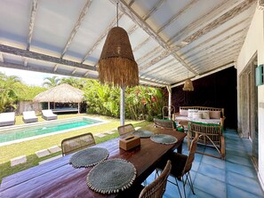 Un área de comedor al aire libre techada con piscina, sillas de descanso y una estructura con techo de paja.