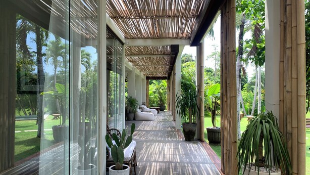 A covered outdoor area with glass walls, a bamboo ceiling, and a pathway lined with potted plants and seating. - Samudra Damai Villas (Pecatu)