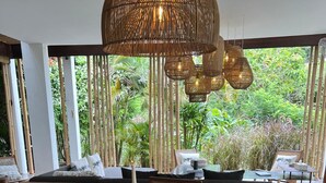 A dining area with a wooden table set for a meal, surrounded by chairs, and a large window offering a view of greenery. - Samudra Damai Villas (Pecatu)