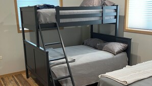 Quarto com beliche, piso de madeira, duas janelas com persianas e uma pequena mesa com um cobertor.