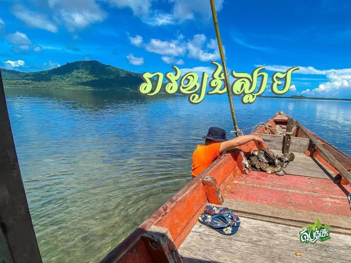 A wooden boat with a person wearing an orange shirt and a hat, a rope tied to the boat's edge, and a clear view of a calm lake with a distant mountain.
