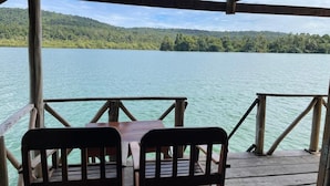 Una terraza de madera con dos sillas y una mesa, con vista a un lago tranquilo y una colina boscosa al fondo.