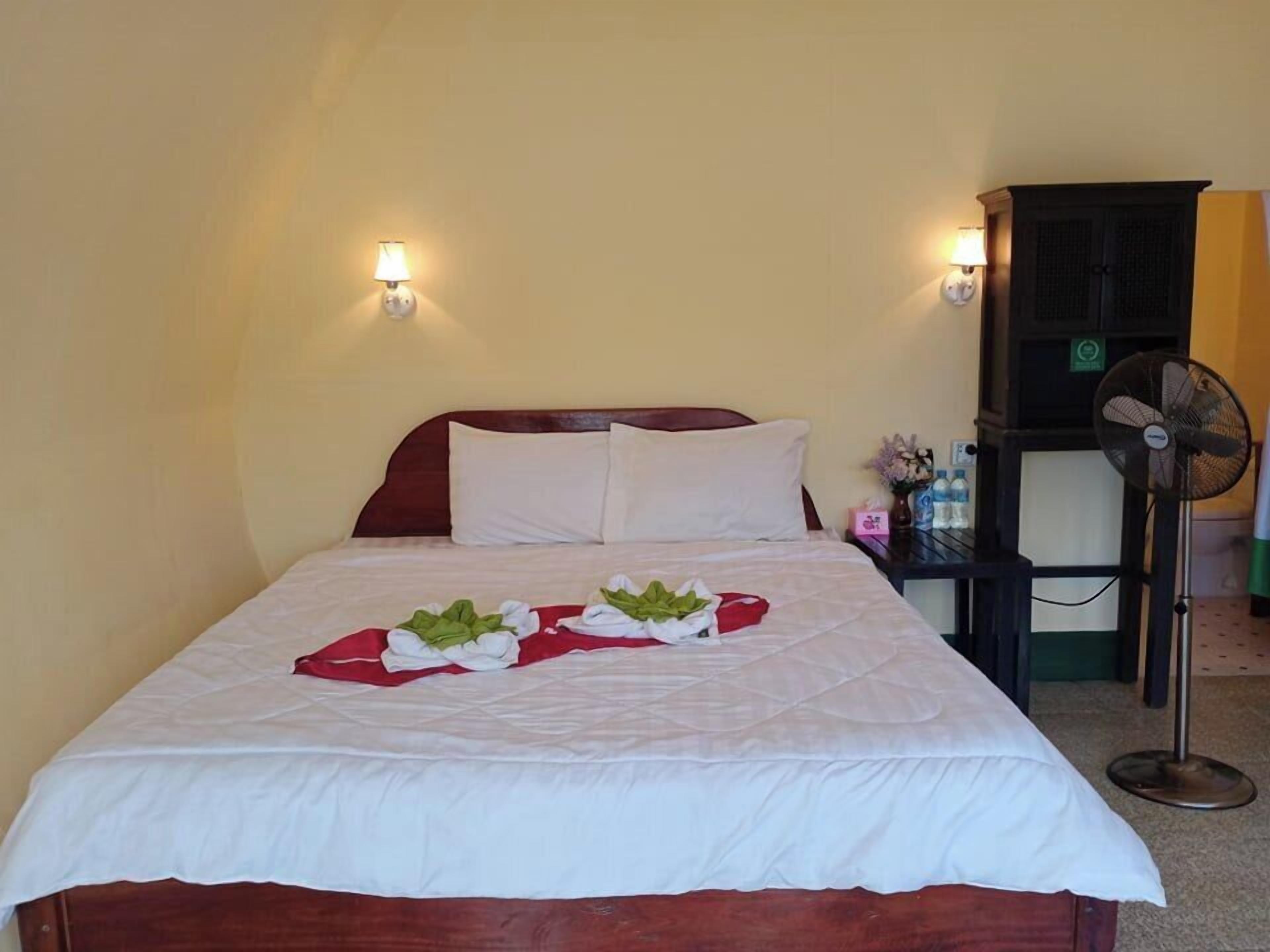 A neatly made bed with a white bedspread and pillows, a bedside table with a lamp and a fan, and a wall-mounted light fixture.