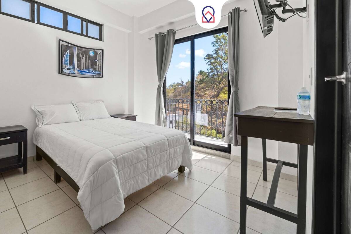 Une chambre à coucher comprenant un lit, une table de chevet, un bureau, une fenêtre avec des rideaux et un tableau encadré accroché au mur.