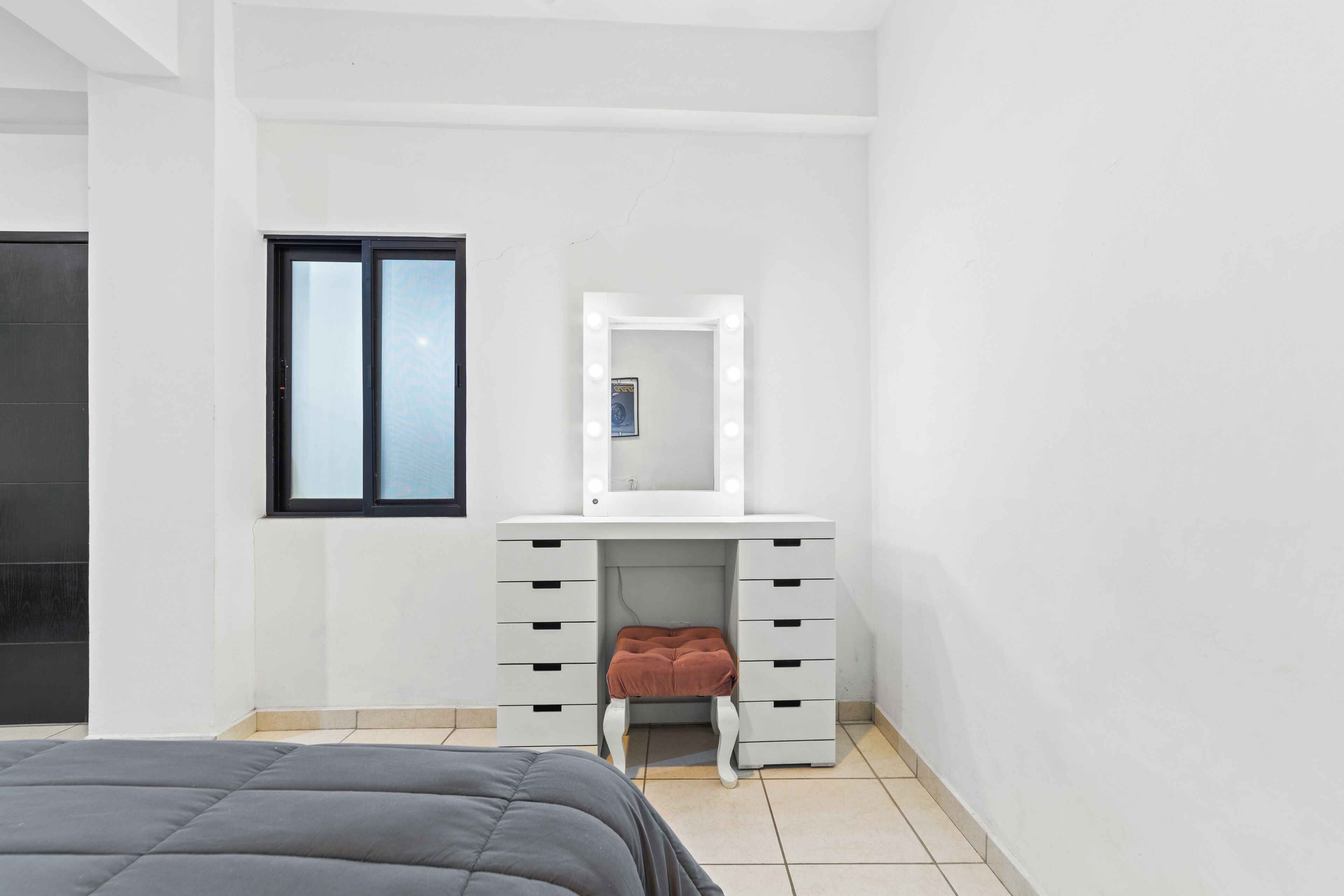 Une chambre à coucher avec un lit, une commode blanche à tiroirs, un miroir avec éclairage intégré et une fenêtre.