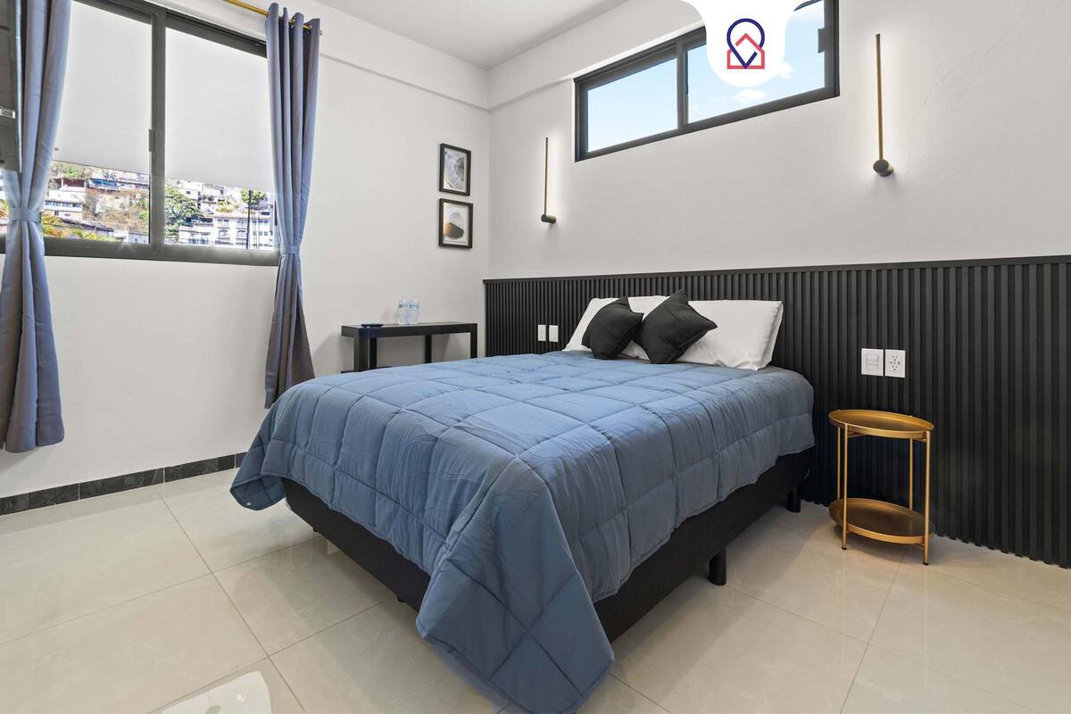 Une chambre moderne avec un grand lit, une petite table de chevet, une fenêtre avec des rideaux et un luminaire fixé au mur.
