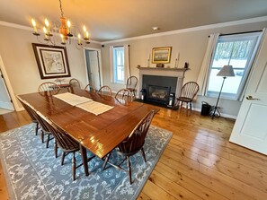 Une salle à manger avec une table en bois, des chaises, un lustre, une cheminée et une grande fenêtre.