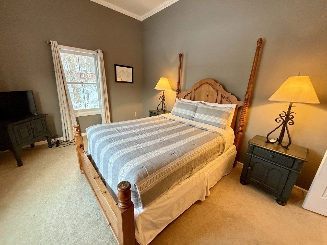 A bedroom with a bed, two bedside tables, a nightstand, a window with curtains, and a wall-mounted lamp.