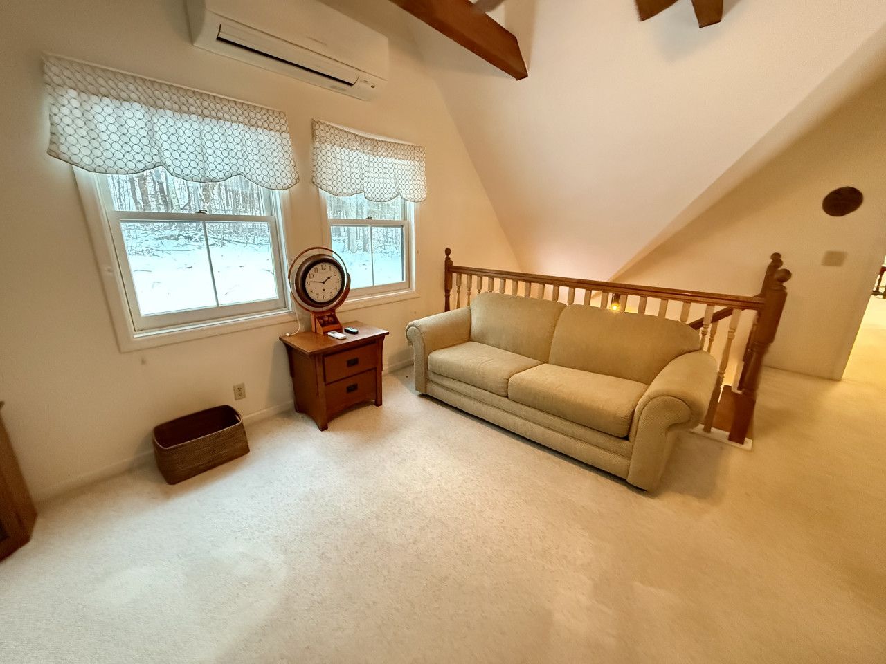 Una habitación acogedora con un sofá beige, una mesita de noche de madera con un reloj y una ventana con un cortinero blanco.