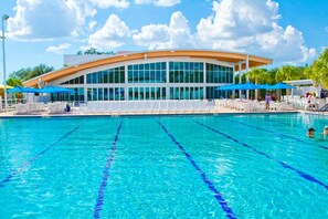 Piscina com água azul cristalina e divisórias de pista, cercada por espreguiçadeiras brancas e guarda-sóis azuis.