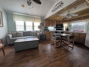 Amplia sala de estar con sofá, reposapiés y mesa de comedor con sillas. Cocina con armarios de madera y electrodomésticos, contigua.