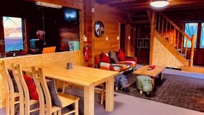 A cozy cabin interior with a wooden dining table, chairs, a sofa with colorful cushions, a small table with a toy pig, and a staircase.