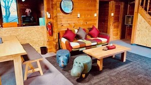 A cozy cabin interior with a wooden wall, a sofa with colorful cushions, a small table, and two animal-shaped stools.
