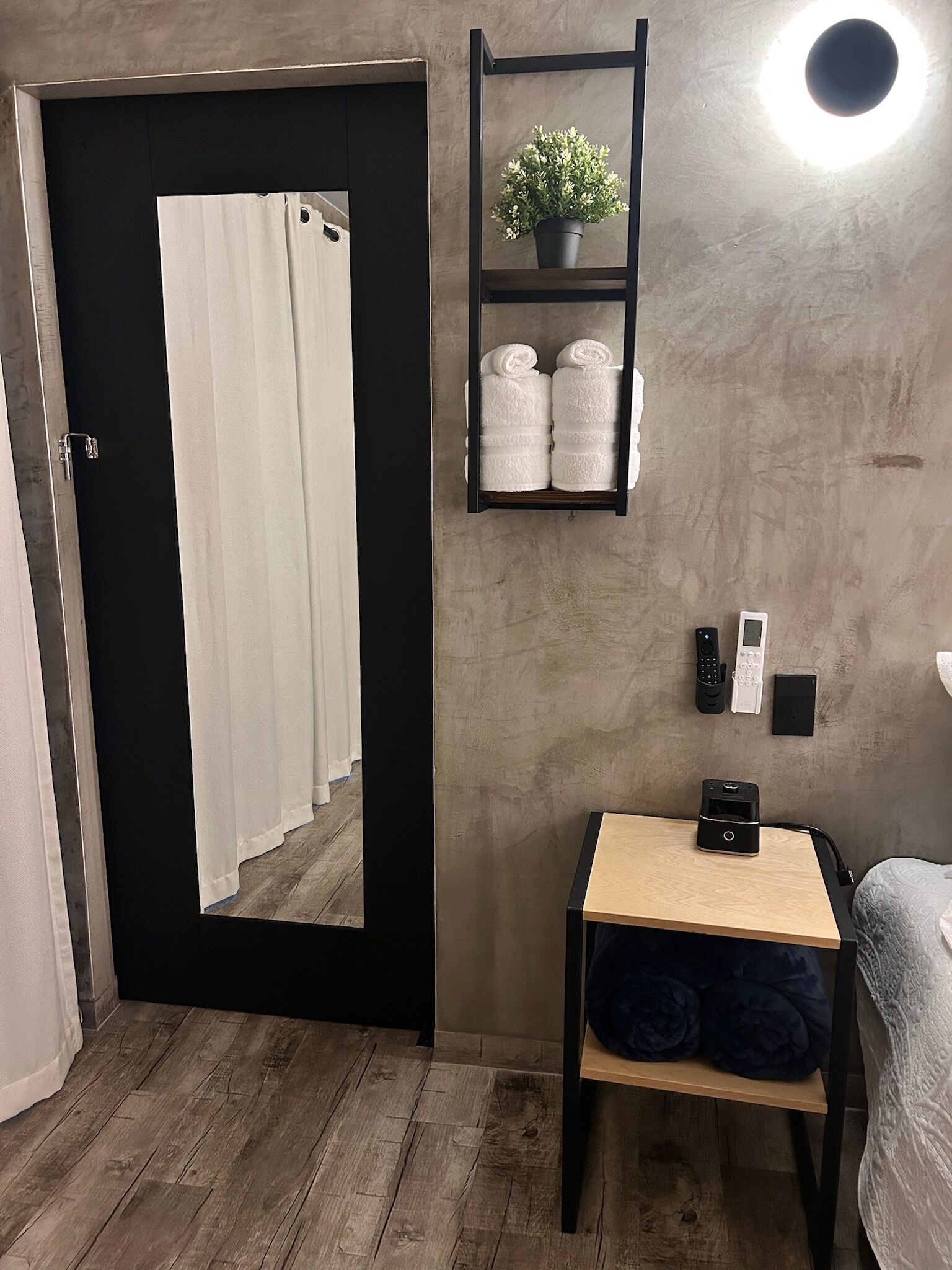 A room with a black door, a small table with towels, a shelf with towels and a plant, and a wall-mounted light.