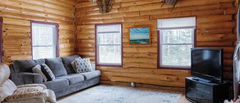 Interior de una cabaña de troncos con un sofá gris, un sillón reclinable, un televisor de pantalla plana y un cuadro en la pared.