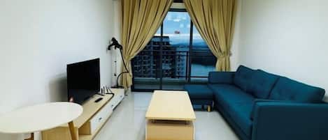 A modern living room with a TV, a sofa, a small table, and a view of buildings through large windows.