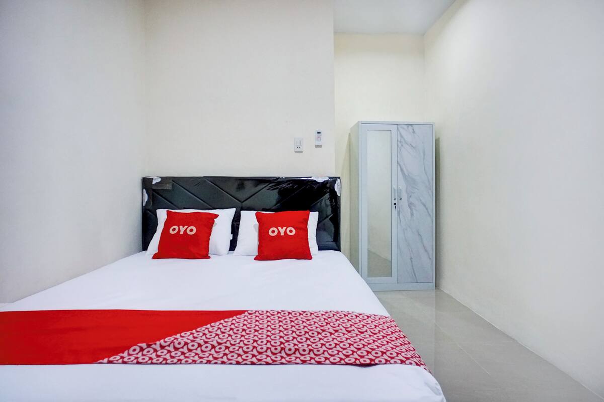 A hotel room with a bed, two red pillows with "OYO" text, a black headboard, a white bedspread, and a red and white patterned blanket.