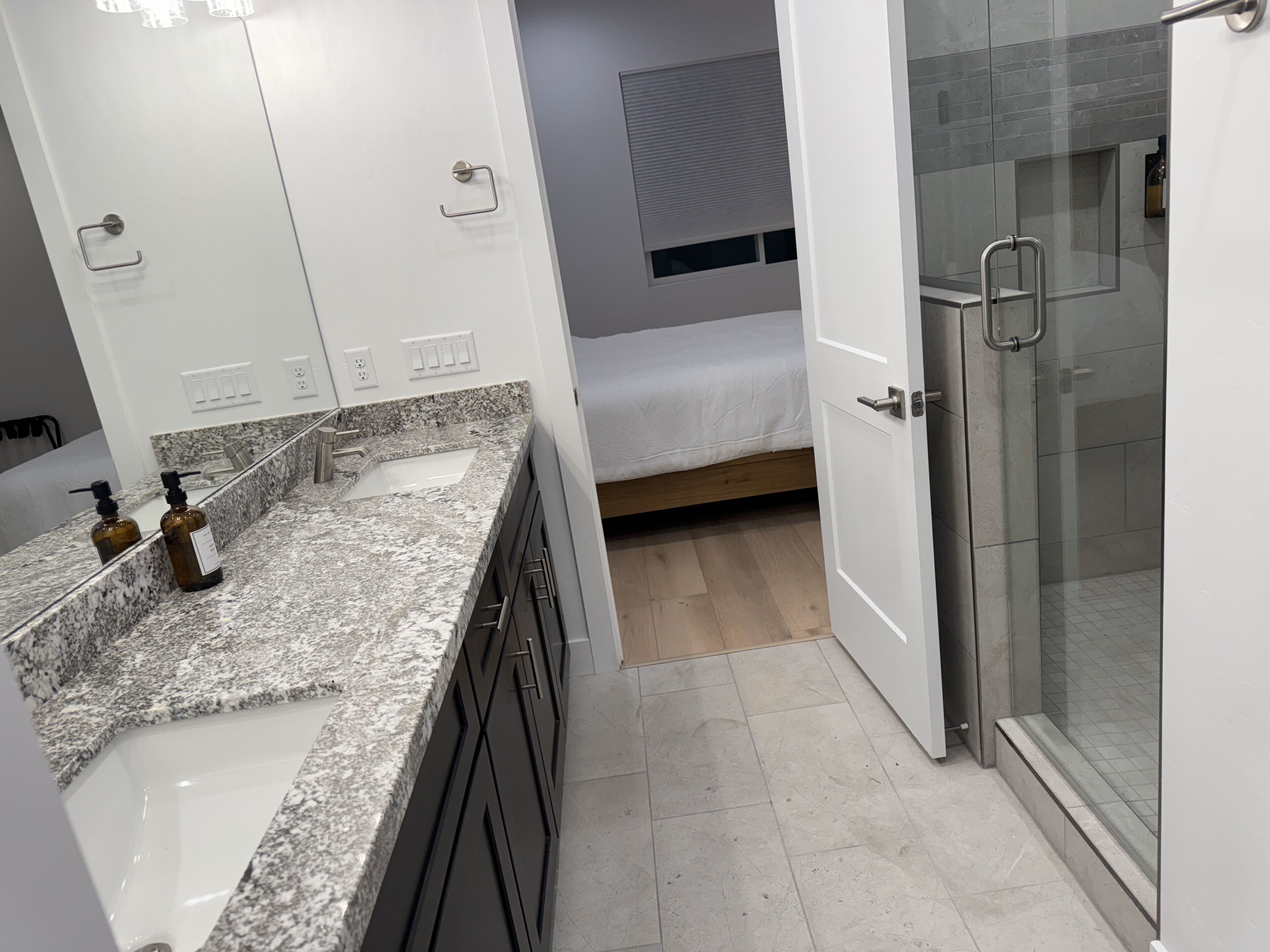A modern bathroom with a granite countertop, a glass shower enclosure, and a bed in the background.