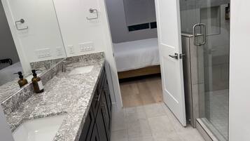 A modern bathroom with a granite countertop, a glass shower enclosure, and a bed in the background.