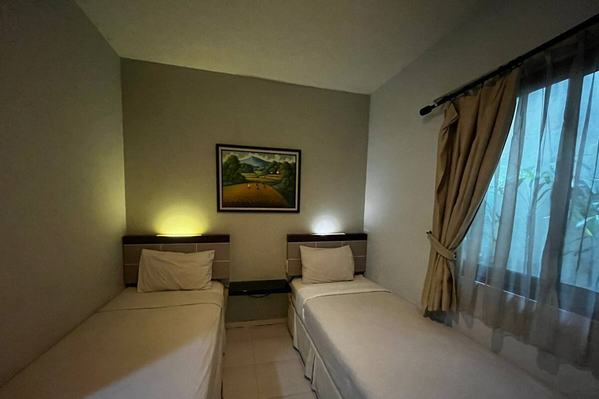 A hotel room with two single beds, a nightstand, a painting on the wall, and a window with curtains.