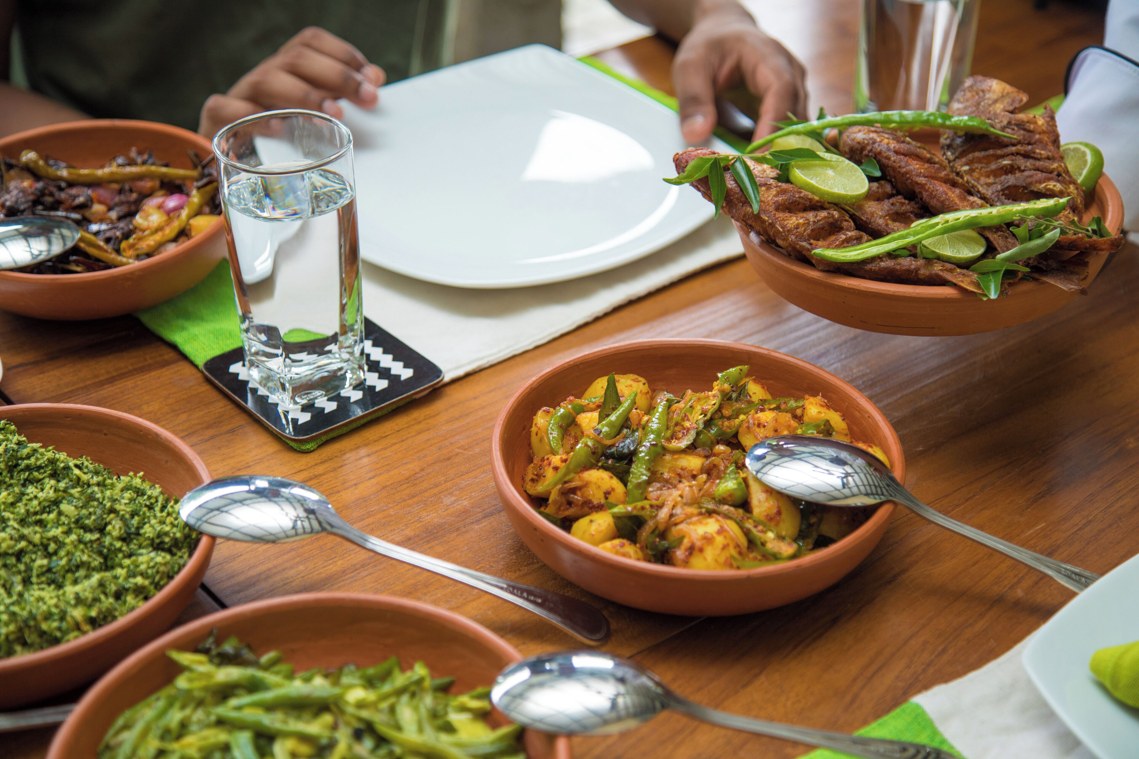 Une table à manger avec différents plats, notamment un bol de légumes verts, un bol de riz jaune et une assiette de poisson frit, garnie de quartiers de citron vert et de piments verts.