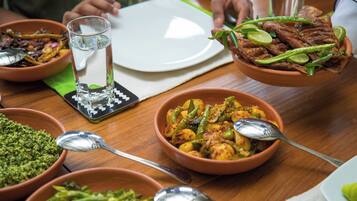 Une table à manger avec différents plats, notamment un bol de légumes verts, un bol de riz jaune et une assiette de poisson frit, garnie de quartiers de citron vert et de piments verts.
