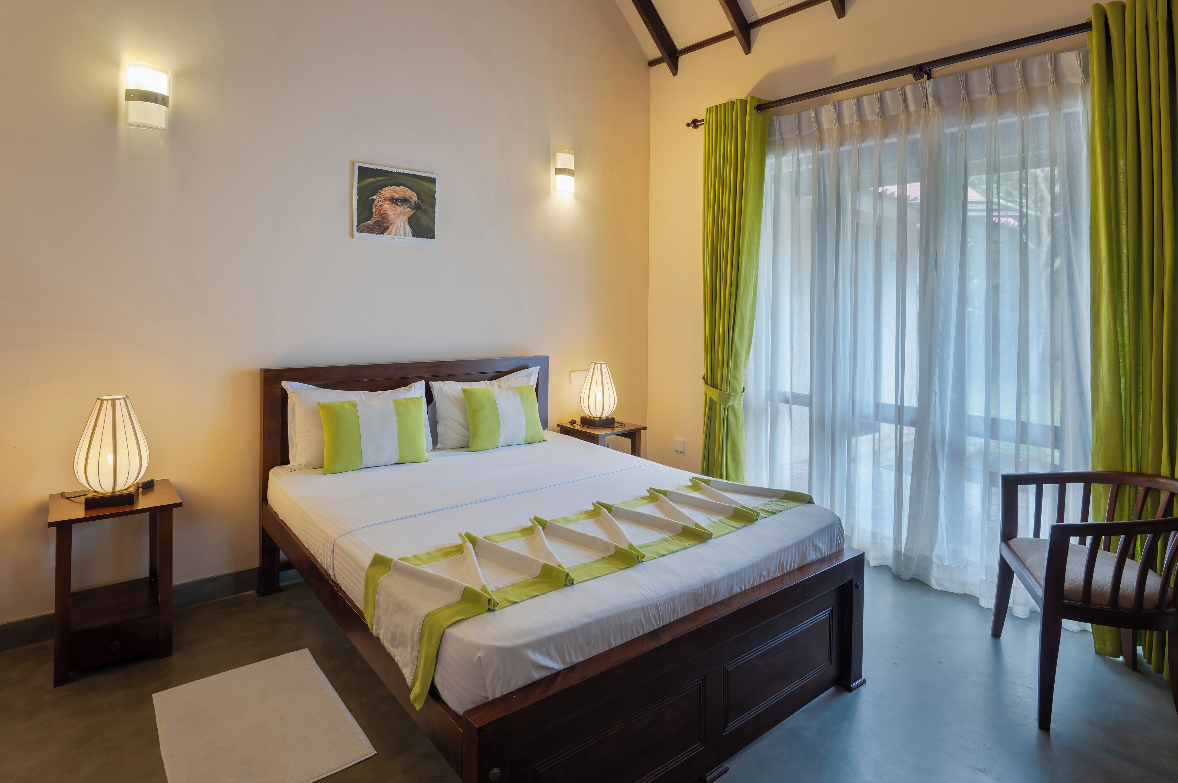 Cama bem arrumada com colcha branca e verde, duas luminárias de cabeceira, um quadro na parede e vista para o exterior através de cortinas leves.
