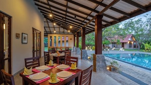 Un'area pranzo coperta con tavoli apparecchiati, una piscina e una vista su una casa sullo sfondo.