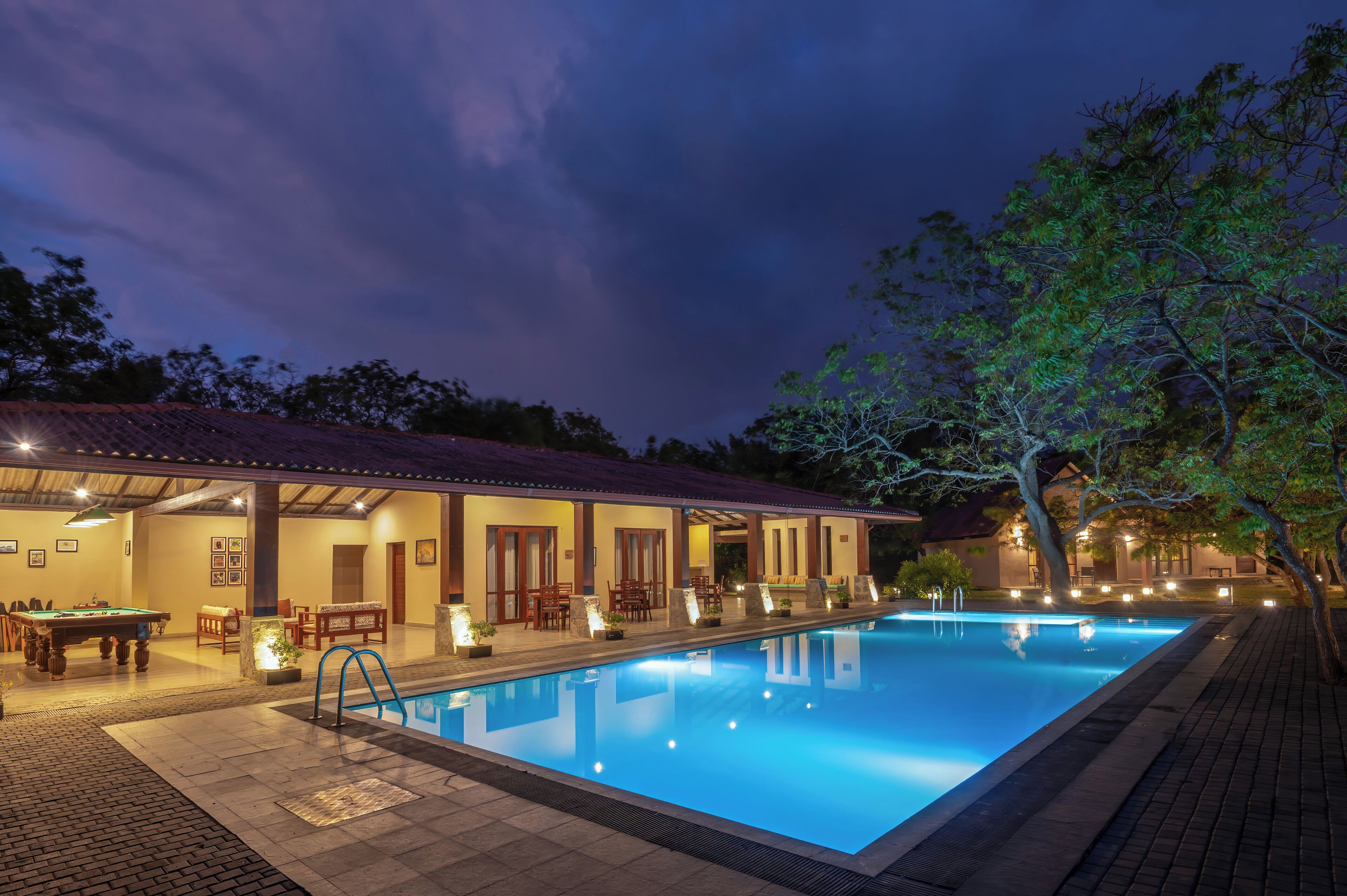 A swimming pool area with a covered seating area, pool table, and outdoor lighting.