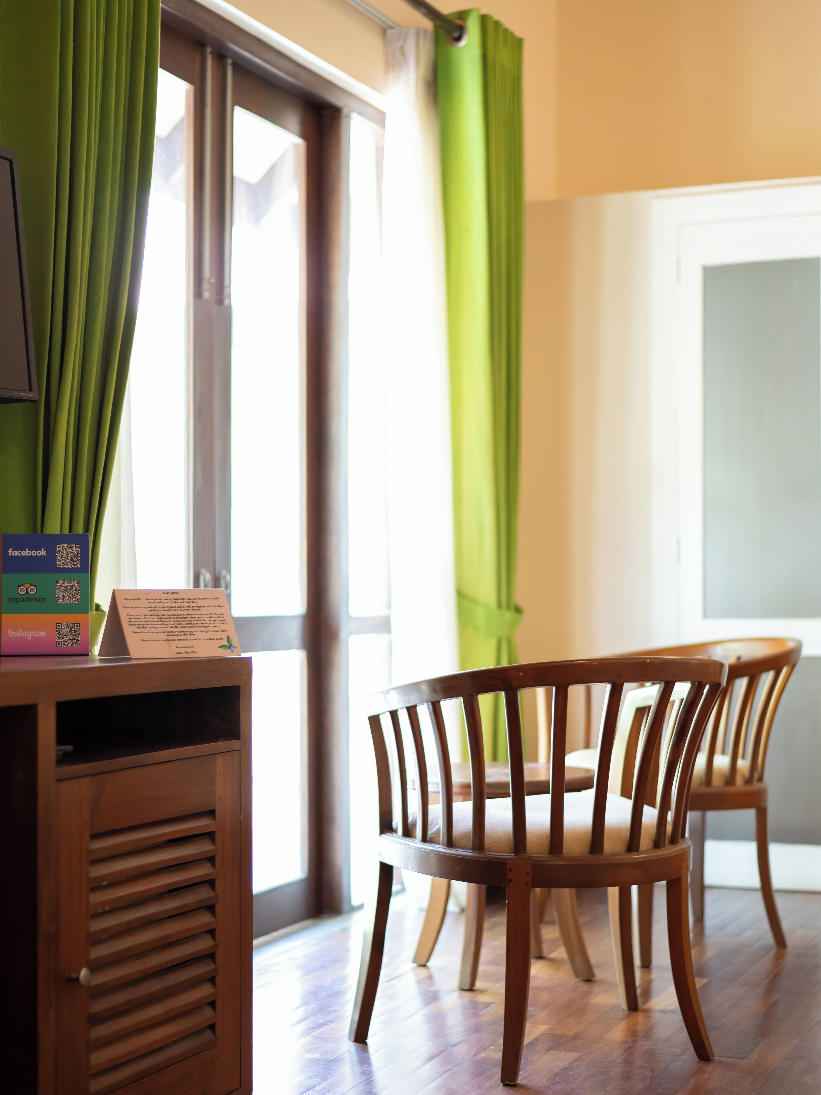 A room with a wooden table and chairs, a window with green curtains, and a sign with QR codes for social media.