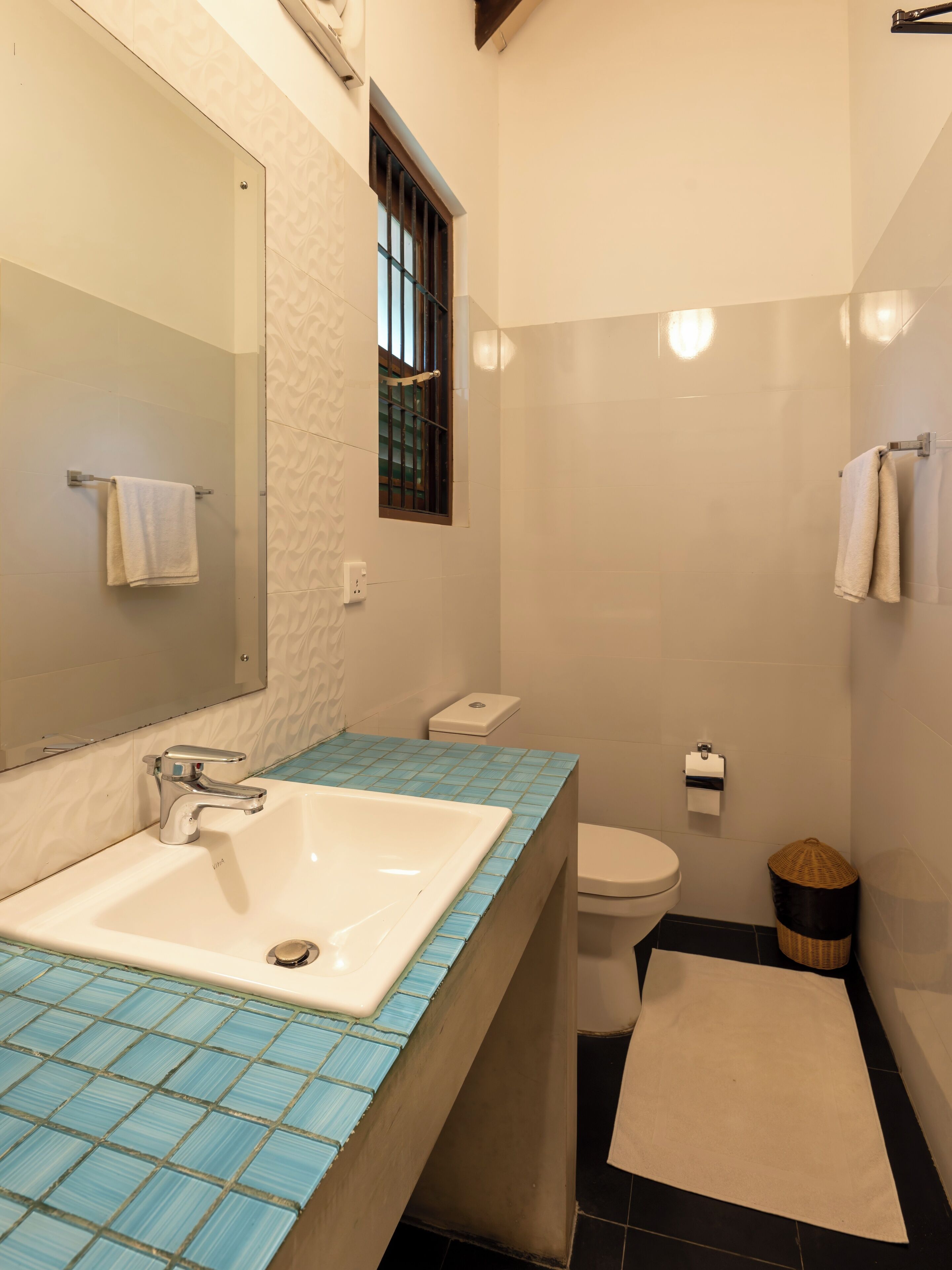 A bathroom with a white sink, a mirror, a window, a toilet, and a small trash bin.