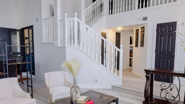 Un amplio espacio de sala con una escalera, un sillón blanco, una mesa de centro con libros y un jarrón decorativo.