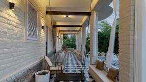 A covered porch with a checkered floor, white brick walls, and wooden furniture. - AbhiRaj Villa (Udaipur)